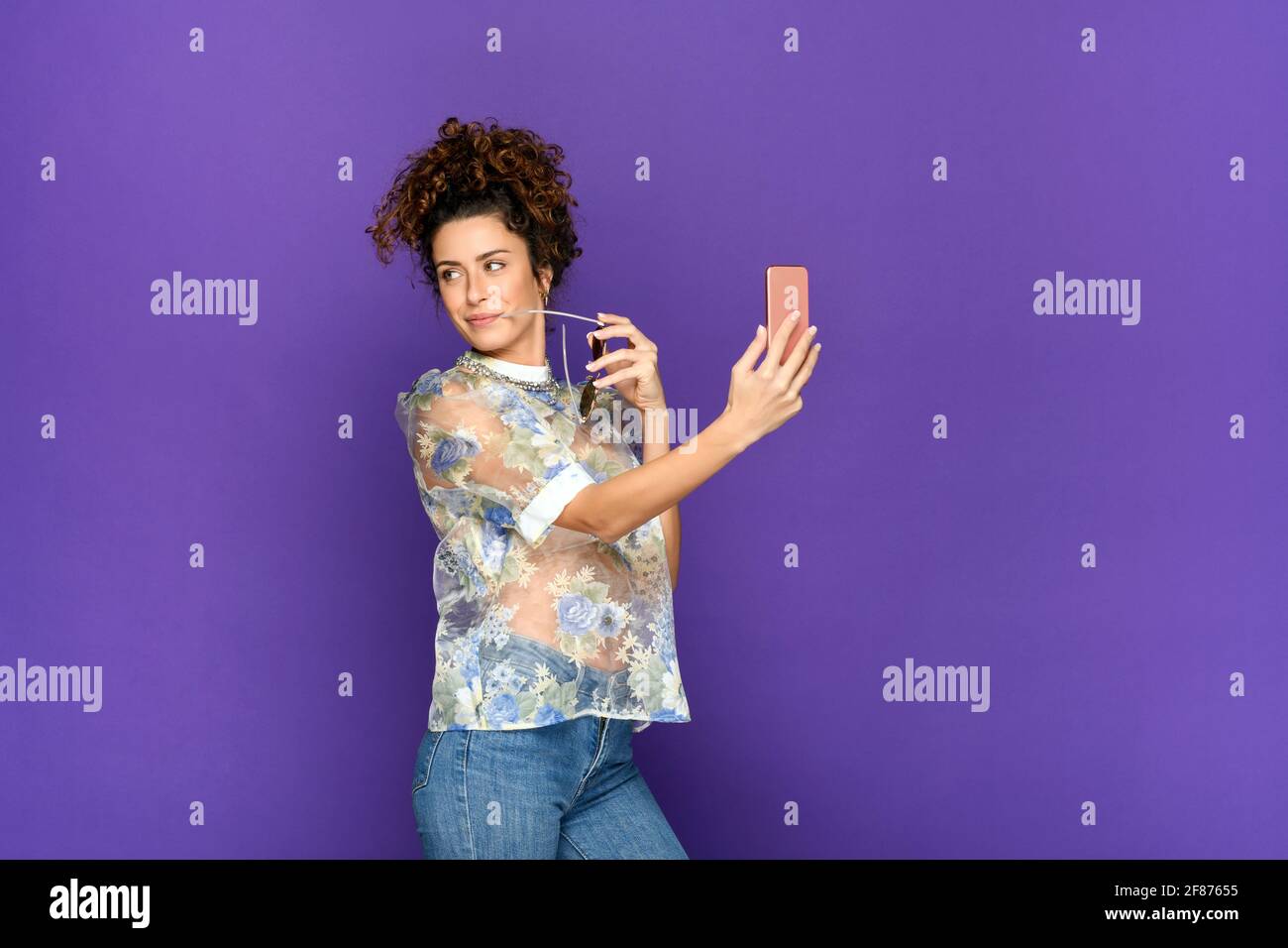 Side view of positive young curly haired brunette in trendy blouse and jeans holding sunglasses and taking selfie on mobile phone while standing again Stock Photo