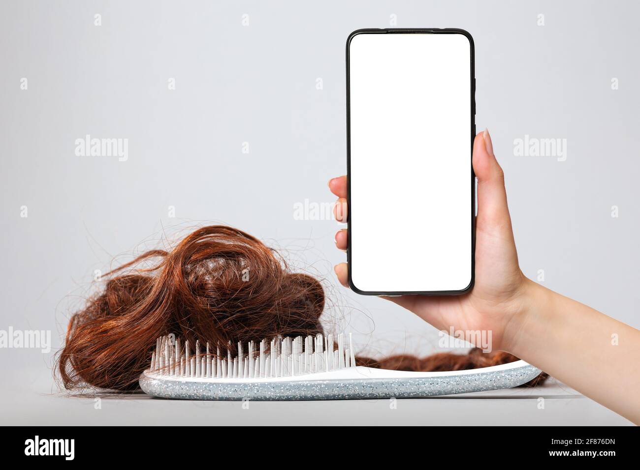 A white comb with false hair. White background. A female's hand holds a smartphone with a white screen. Mock up. The concept of buying and selling hai Stock Photo