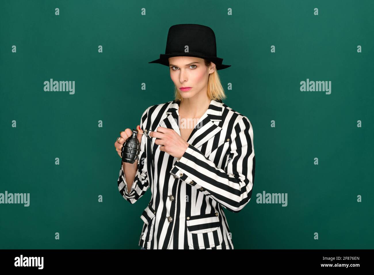 Serious female in stylish striped jacket and hat looking at camera and pulling grenade pin against green background Stock Photo