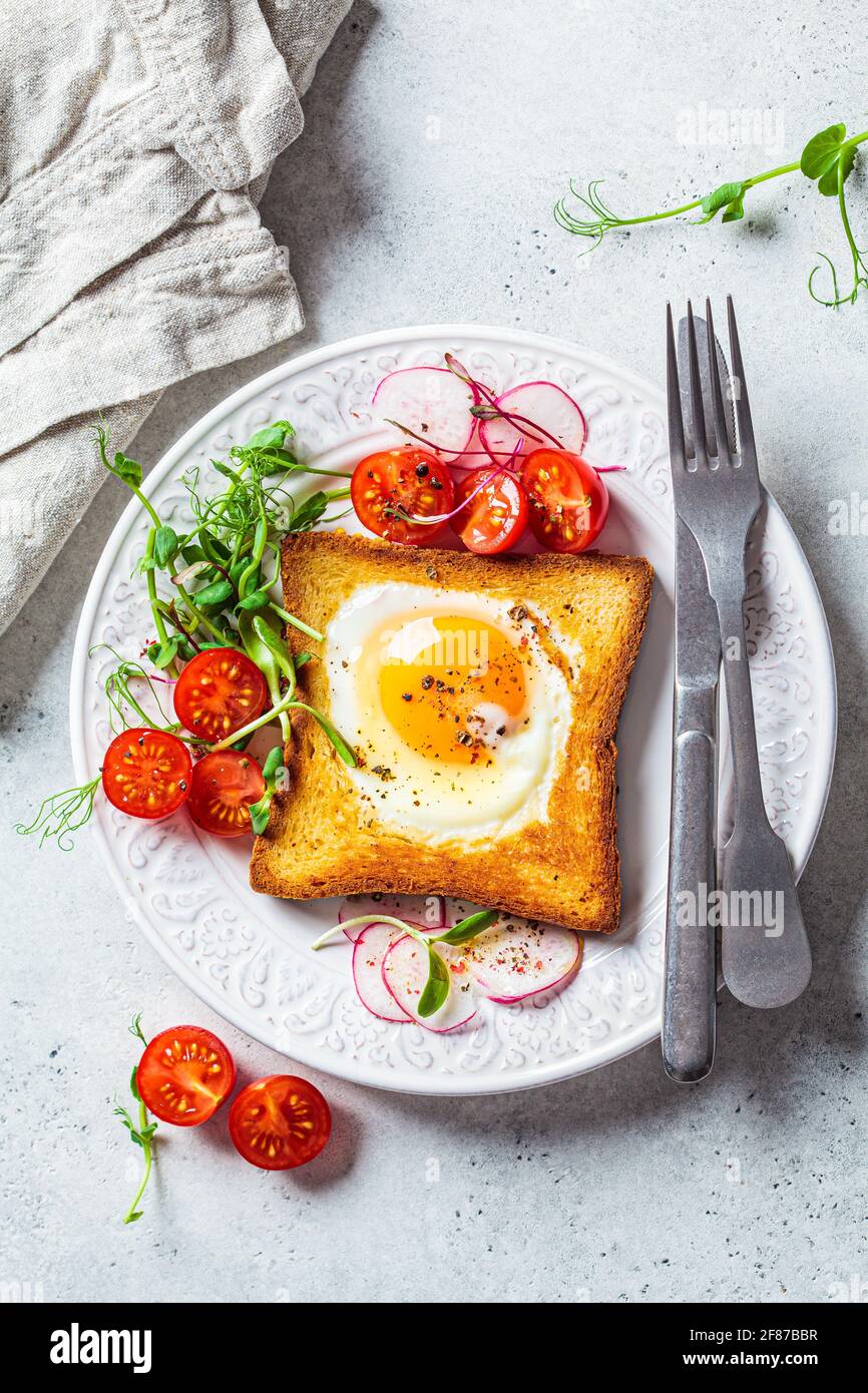 Fried egg in bread toast with tomatoes, radishes and sprouts on a white plate, gray background. Healthy breakfast concept. Stock Photo