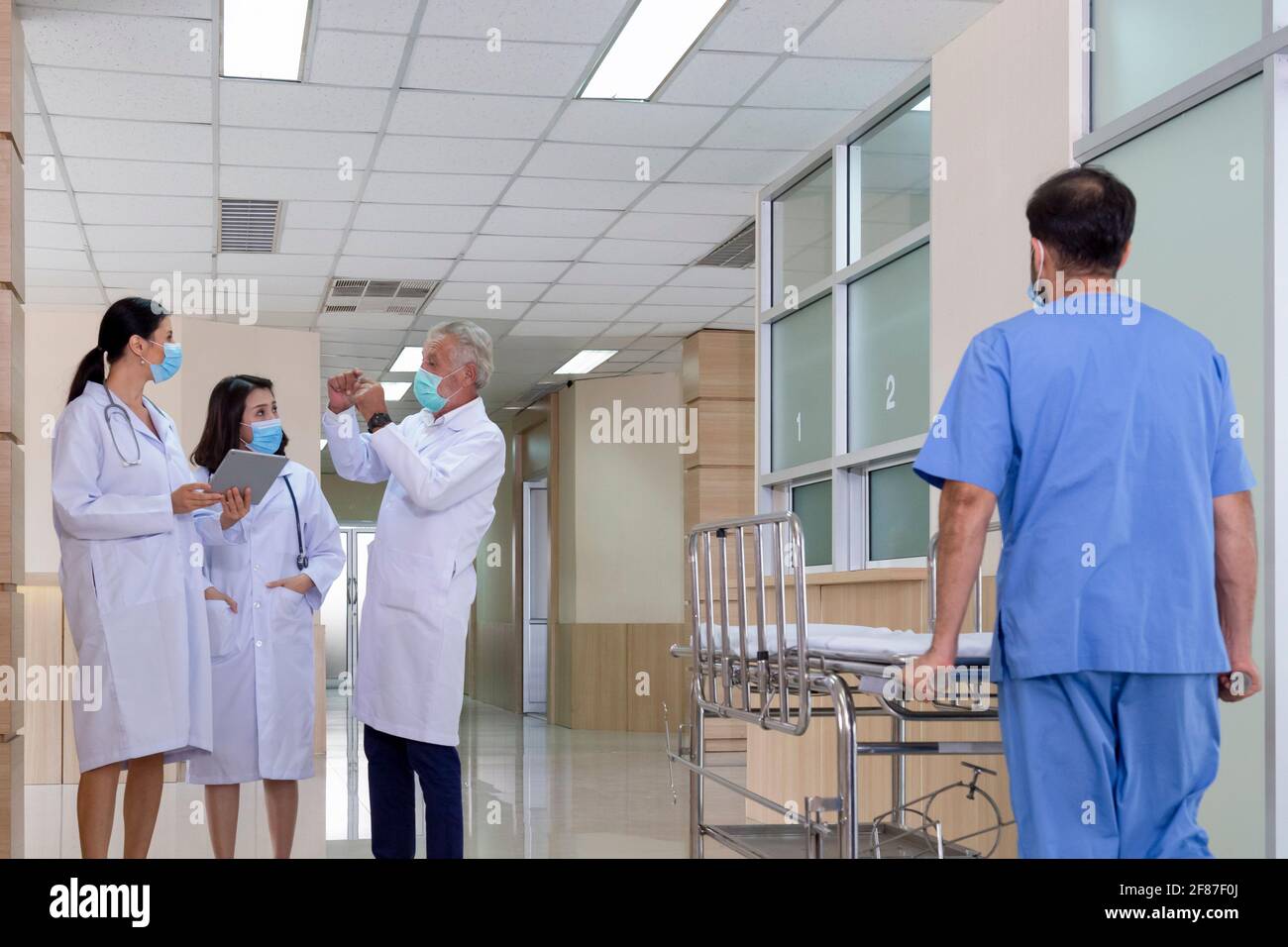 senior professional doctor consult and discuss with woman doctors in serious tumor and cancer surgery case while male nurse pushing stretcher gurney b Stock Photo