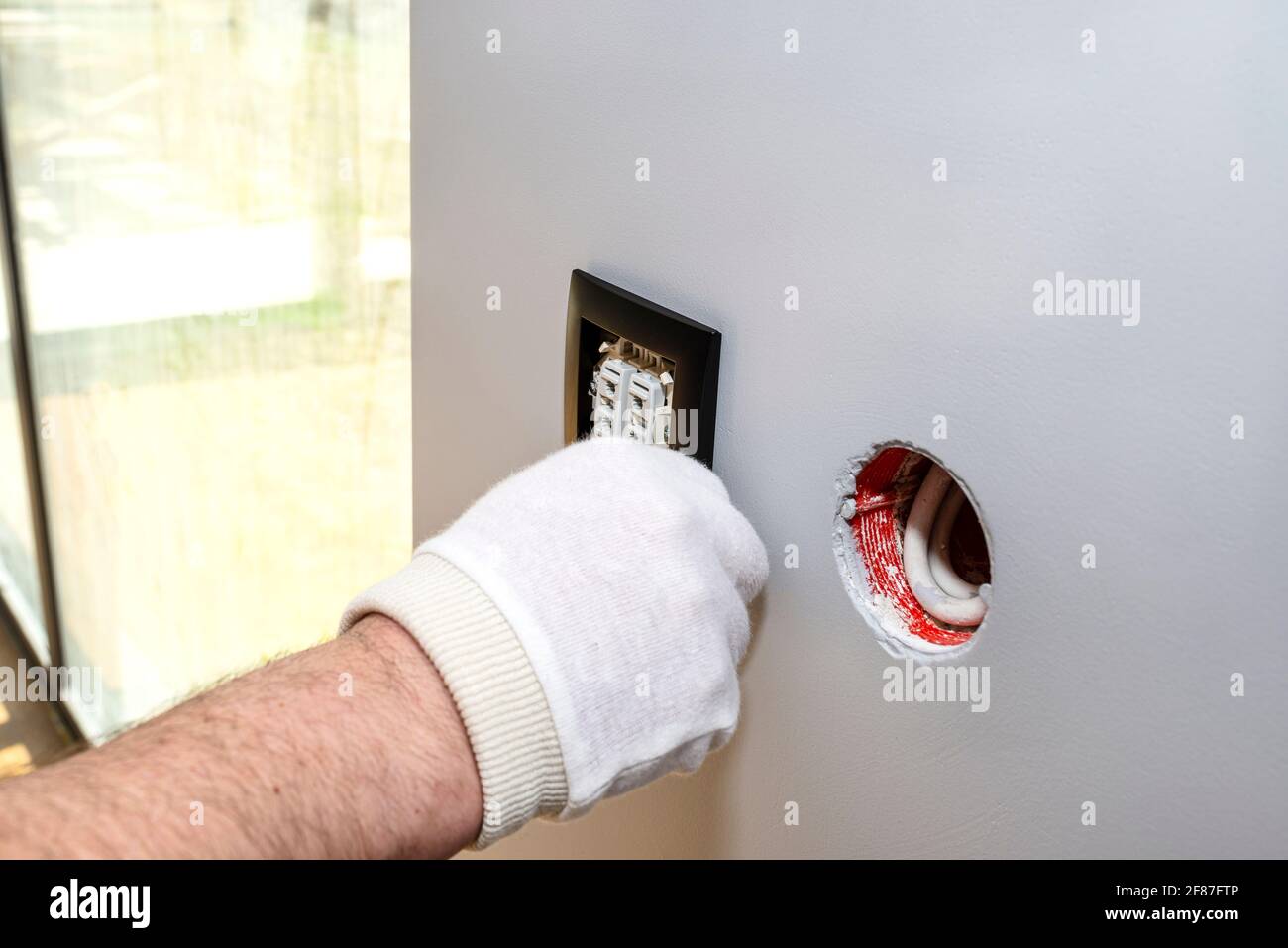 An electrician mounts the black frame of the roller shutter switch up and down with white gloves in the room by the window. Stock Photo