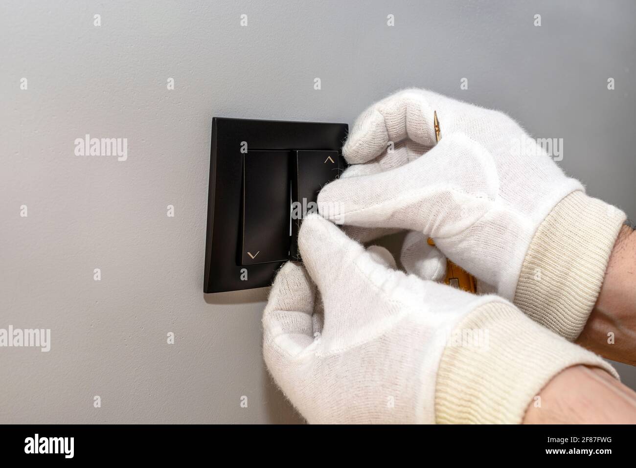An electrician mounts the black button of the roller shutter switch up and down with white gloves in the room by the window. Stock Photo