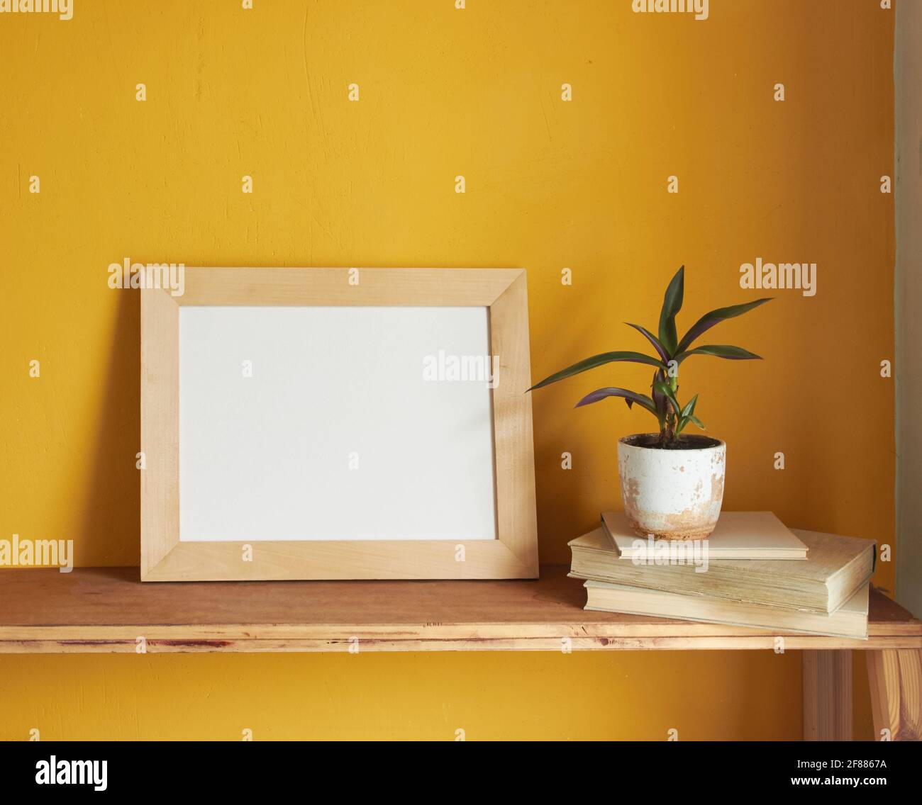 Wooden picture frame mockup. Flowerpot on a pile of books on an old wooden shelf. Composition on a yellow wall background Stock Photo