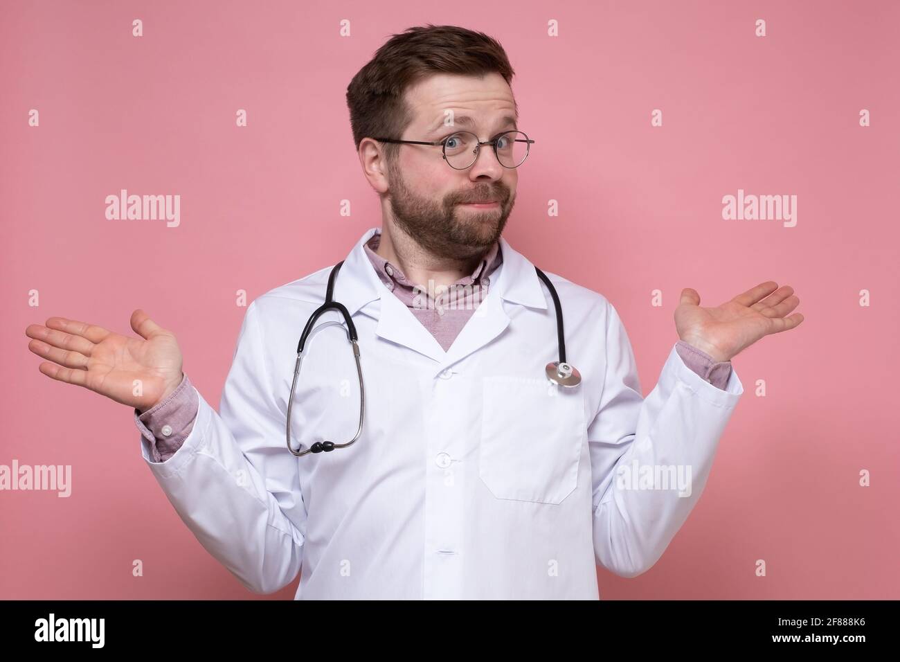 Surprised doctor with a stethoscope around neck innocently spreads hands and looks at the camera. Pink background. Stock Photo