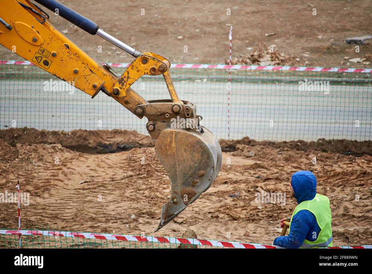 Astrakhan, Russia - 02.15.2021: A worker in a blue jacket and green signal vest he watches the backhoe bucket dig into the ground Stock Photo