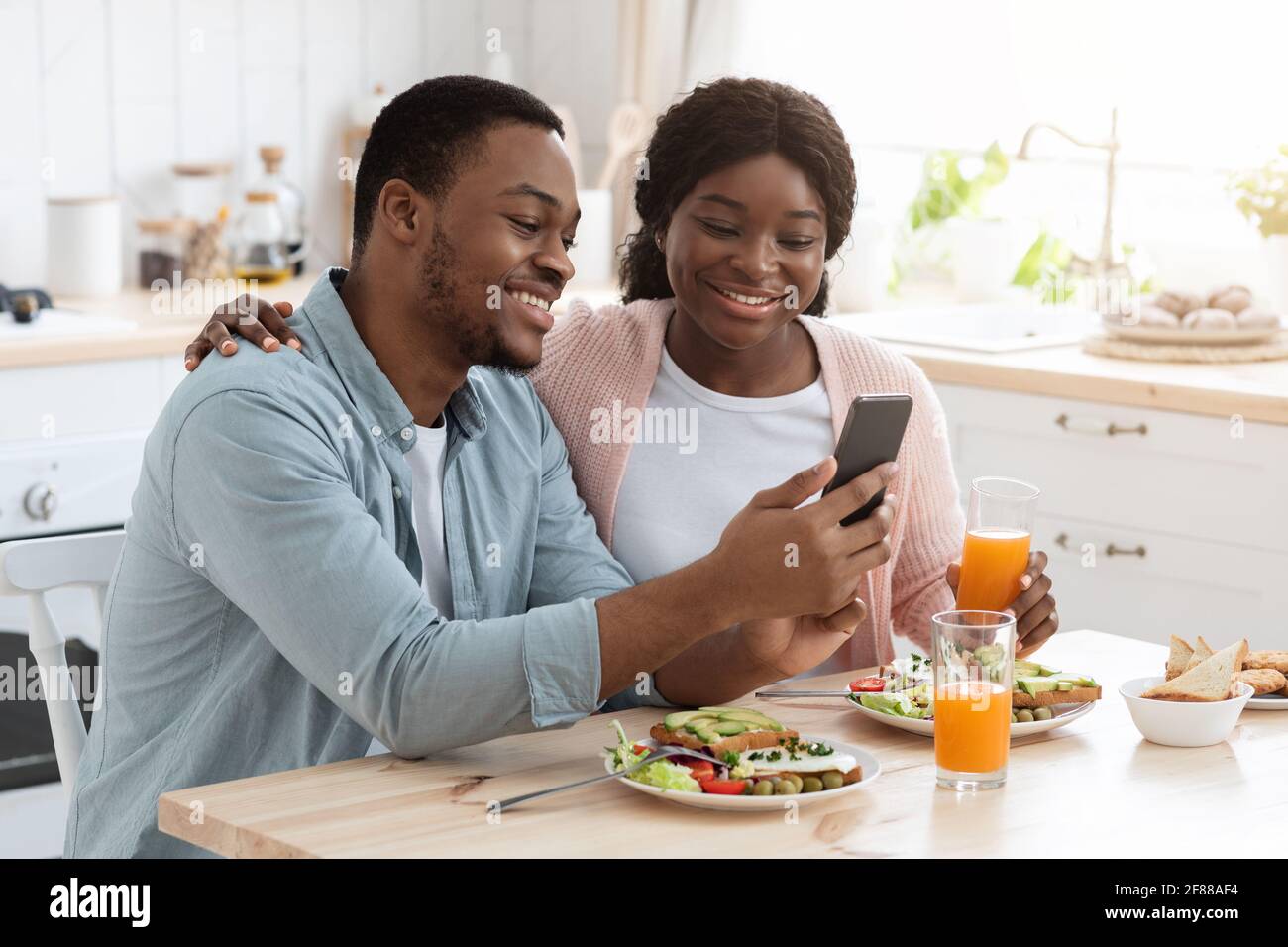 Happy African American Spouses Using Smartphone While Having Breakfast In Kitchen Stock Photo
