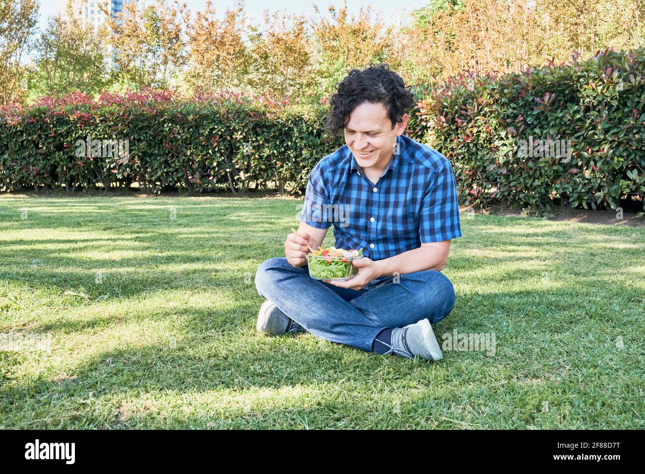 Smiling young man sitting on the grass eating a fresh salad. Healthy living and eating concepts. Stock Photo