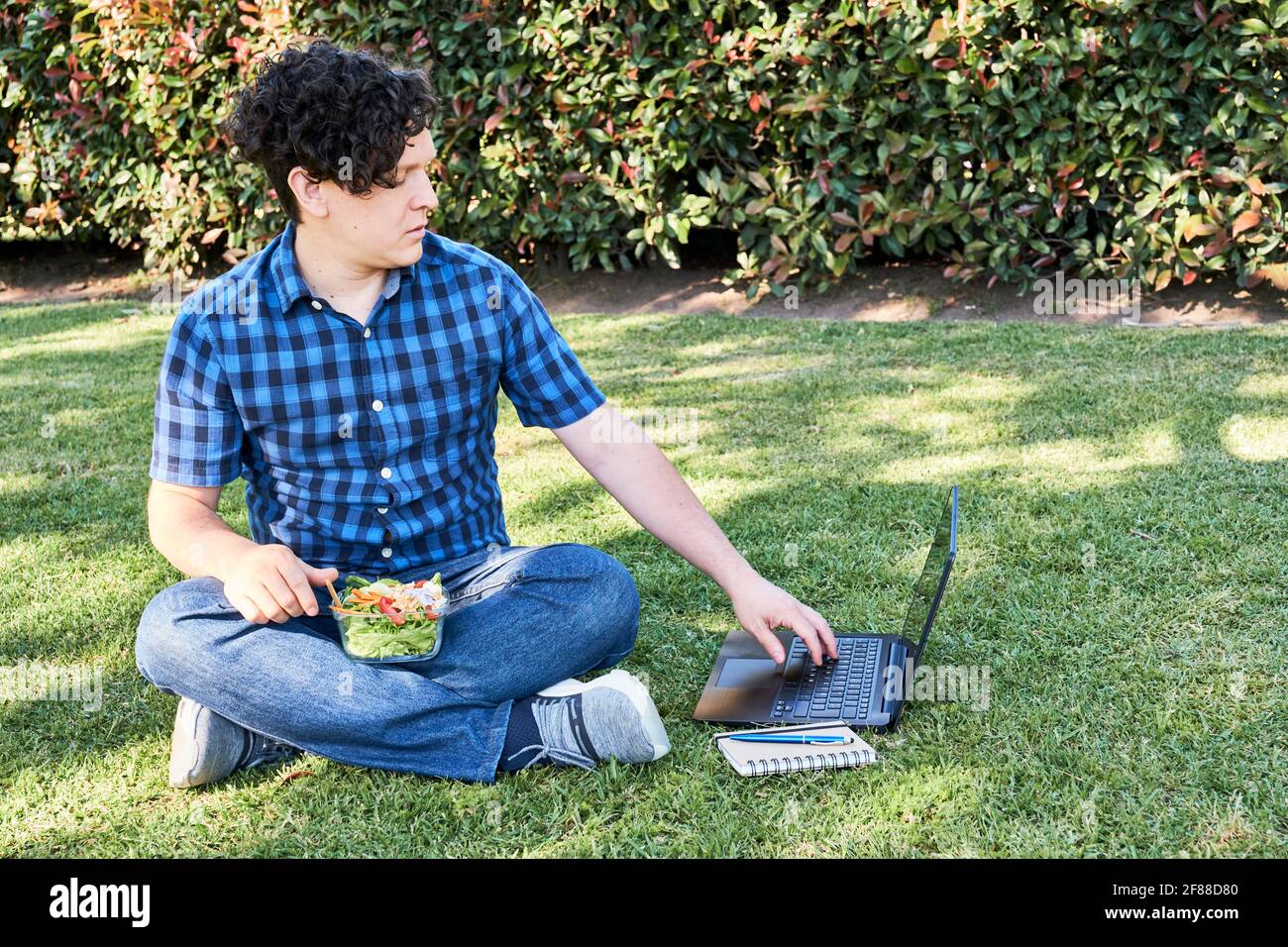 Young man sitting in the grass eating a fresh salad while working or studying on his laptop. Concepts of working outdoors and healthy living and eatin Stock Photo