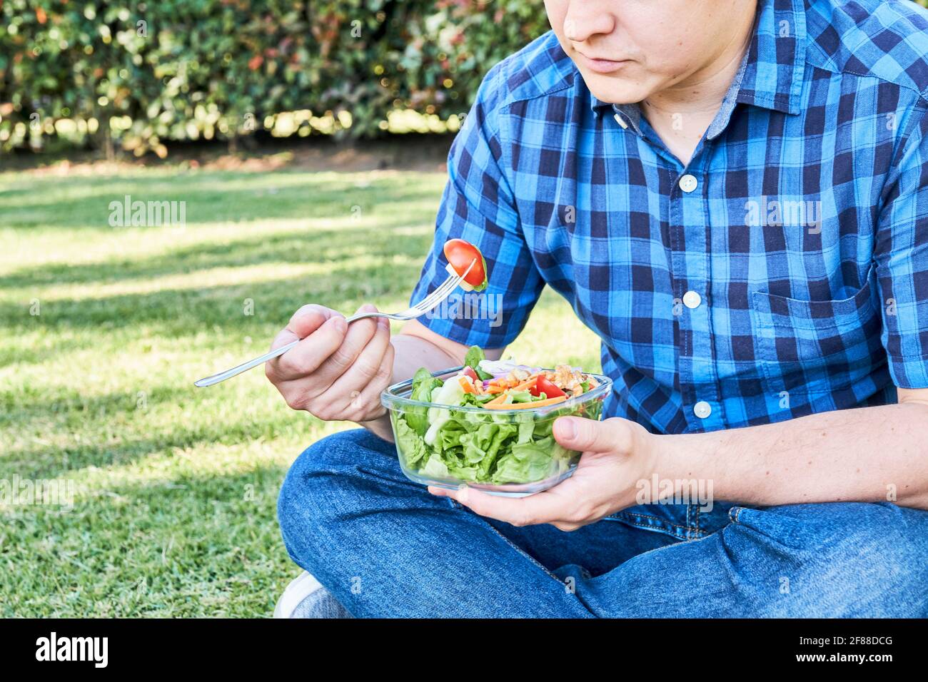 Unrecognizable young man sitting on the grass eating a fresh and colorful salad held in a glass bowl. Healthy living and eating concepts. Close up ima Stock Photo