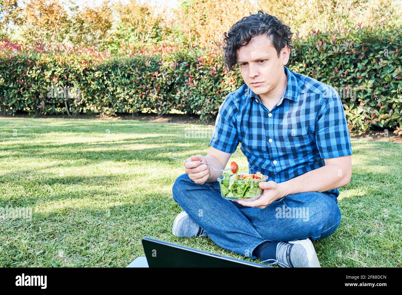 Young man sitting in the grass eating a fresh salad during a break, while working or studying on his laptop. Concepts of working outdoors and healthy Stock Photo