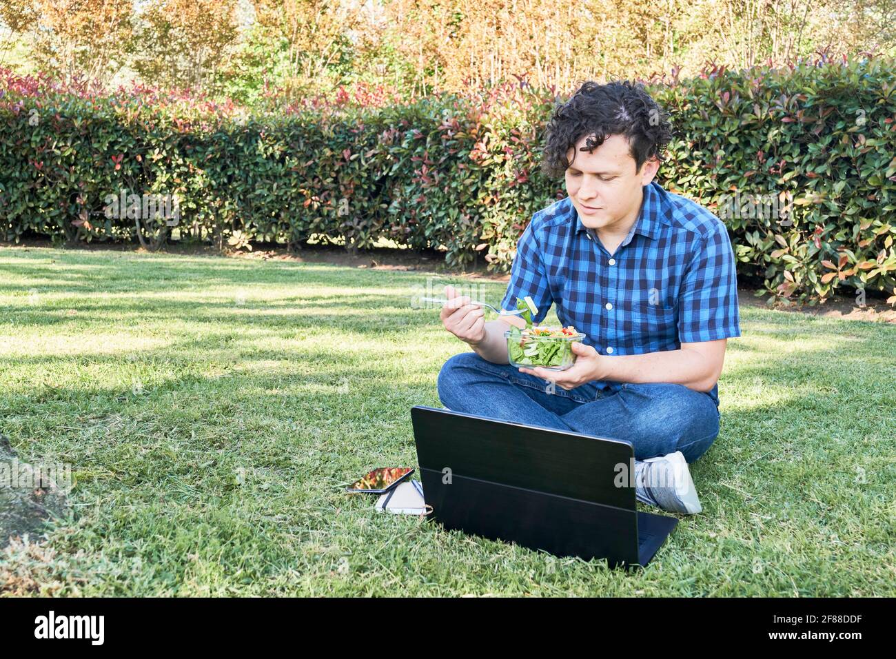 Young man sitting in the grass eating a fresh salad during a break, while working or studying on his laptop. Concepts of working outdoors and healthy Stock Photo