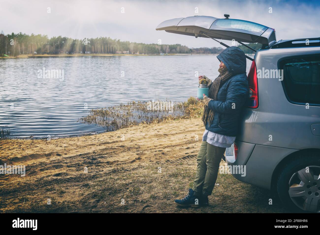 Woman is eating a hot dish from a thermos during a hike, leaning against a car by a lake, with a forest in the background Stock Photo