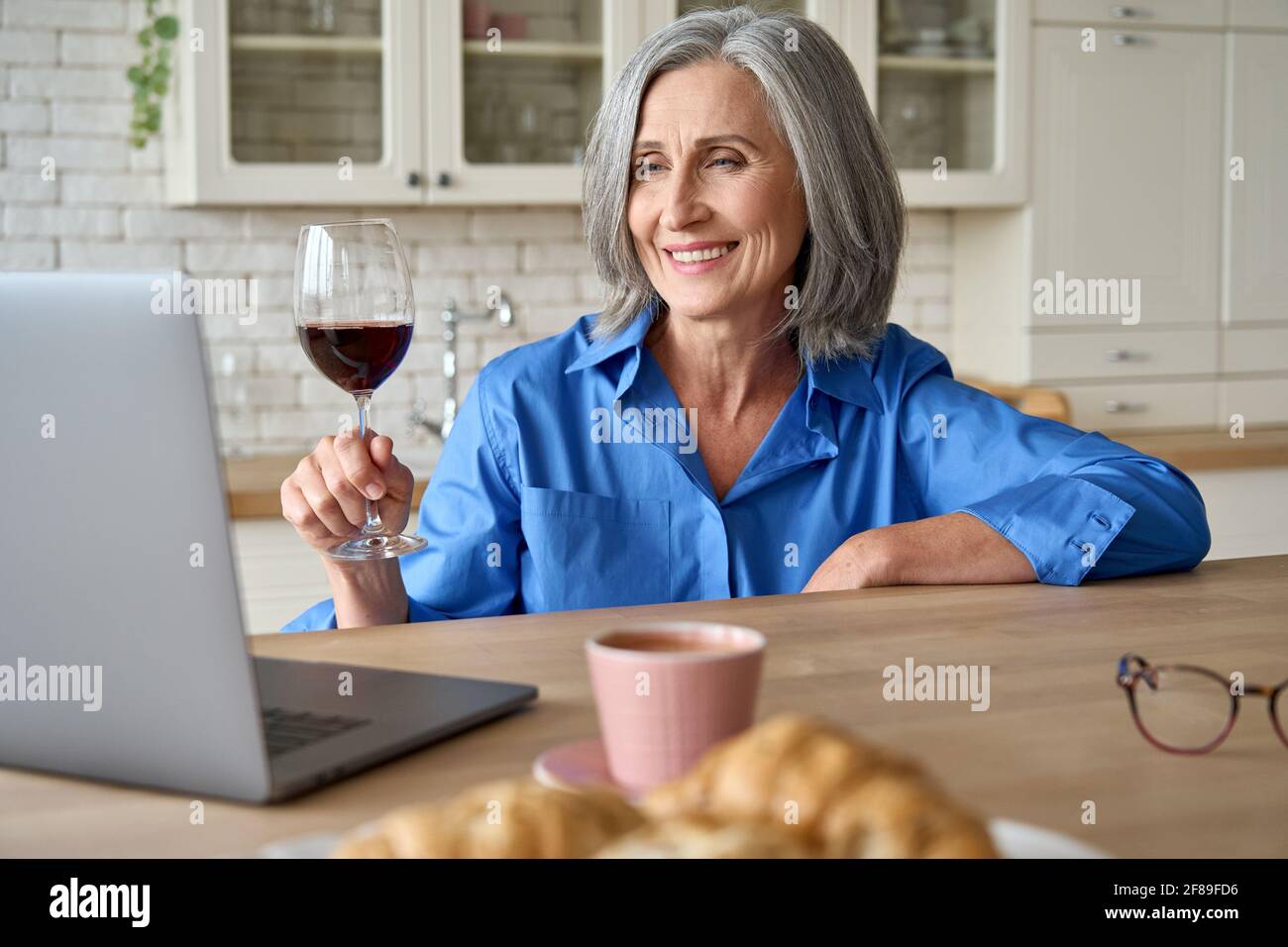 Middle age 60s woman sitting at home drinking wine online with family. Stock Photo