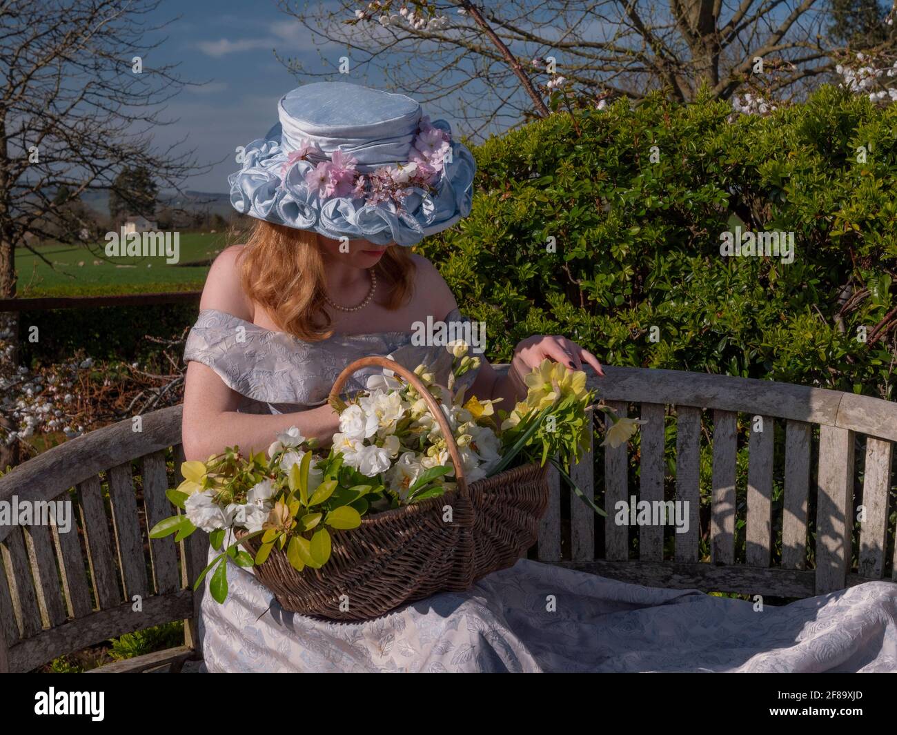Young woman sitting on the bench on the viewing platform dressed in the Easter bonnet with off shoulder dress. The green field in the backdround. Stock Photo