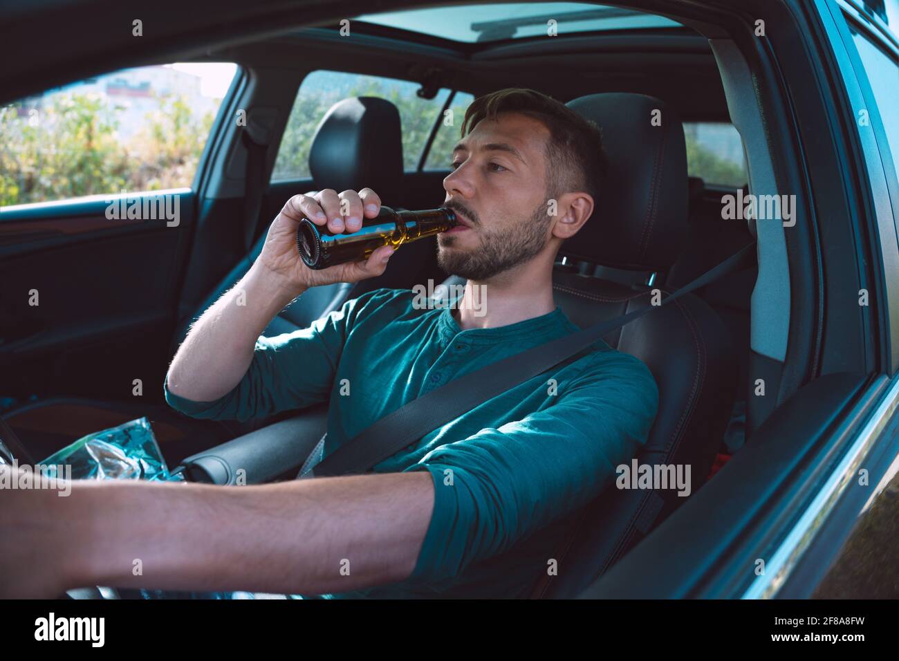 Drunk driver. Young man drinking beer while driving a car. Driver under alcohol influence. Dangerous driving concept. Stock Photo