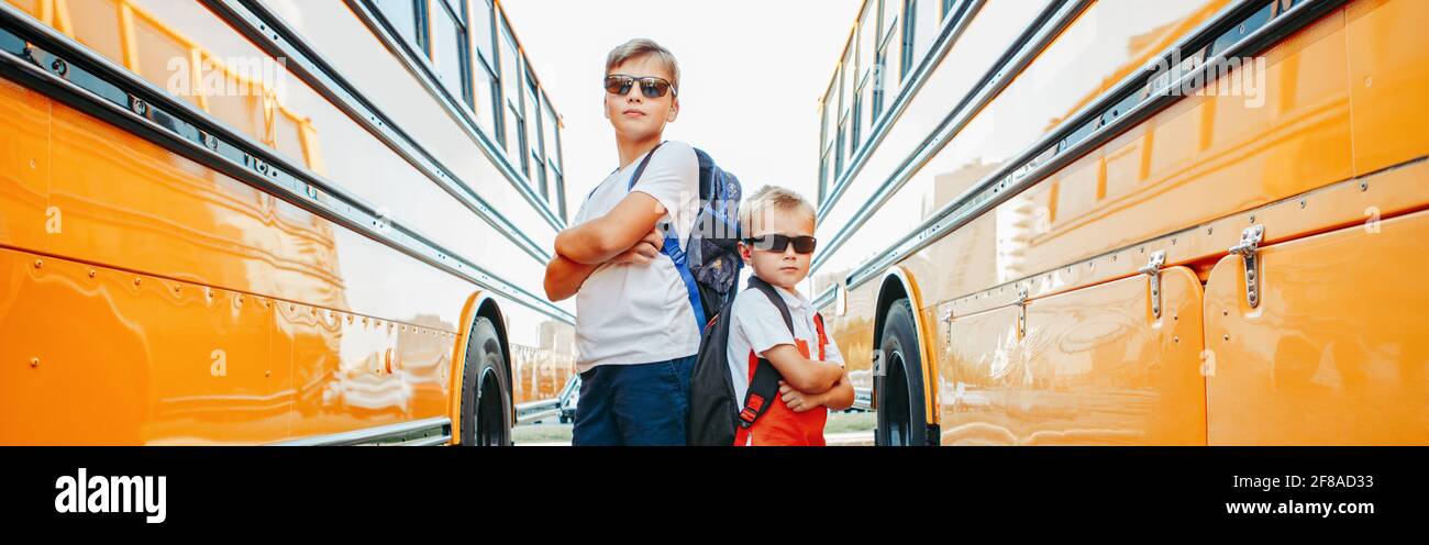 Caucasian brothers students near yellow school bus. Cool kids in sunglasses going back to school in September. Education system and learning. Support Stock Photo