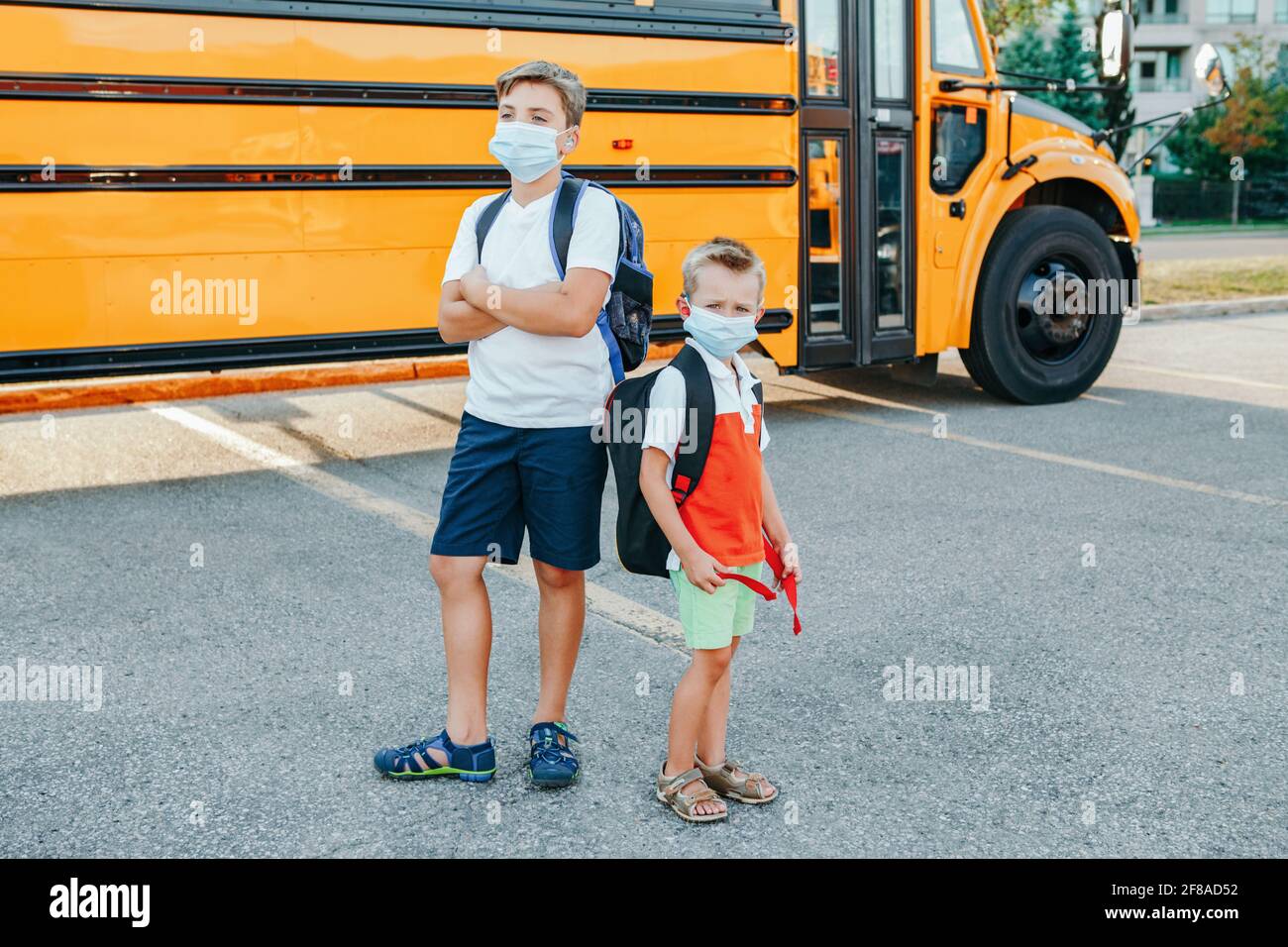 Brothers students in face masks standing by yellow school bus outdoor. Kids with personal protective equipment on faces. Education and back to school Stock Photo