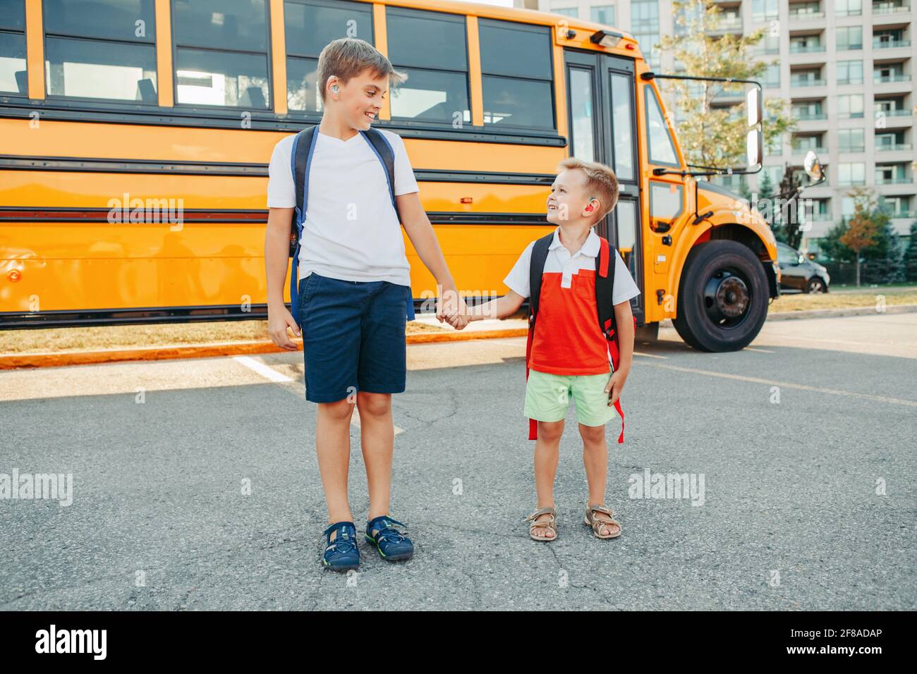 Happy Caucasian brothers students with hearing aids by yellow school bus. Smiling kids with disabilities holding hands. Back to school in September. E Stock Photo