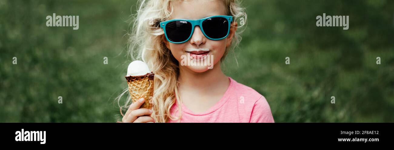 Cute funny adorable girl in sunglasses with dirty nose and moustaches eating ice cream from waffle cone. Happy cool hipster child eating tasty food Stock Photo