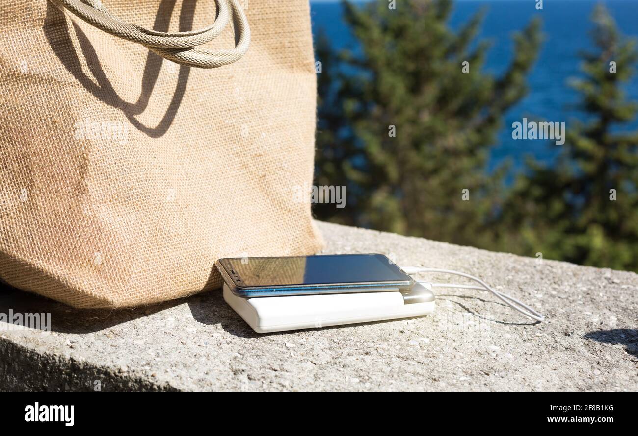 Power bank charges a smartphone on the sea front near the beach bag mock up. A portable travel charger. Stock Photo