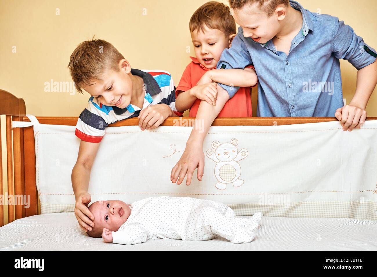 Happy elder brothers look carefully and touch gently lovely baby sibling wearing soft white body and lying in wooden crib Stock Photo