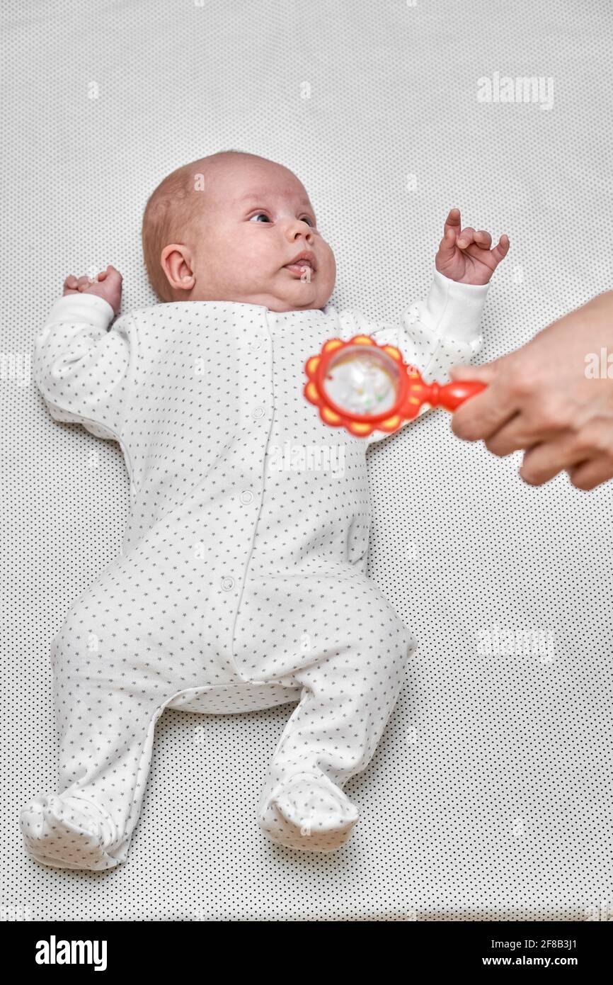 Sweet little newborn baby with tiny fingers wearing white body lies and looks at mother holding orange maraca close view Stock Photo