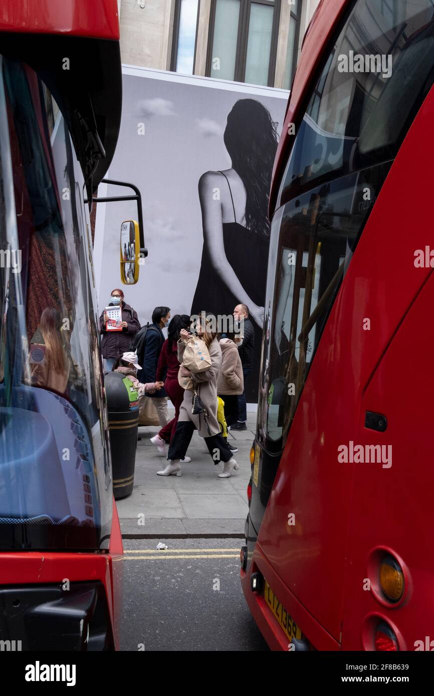 On the day that the UK government eased Covid restrictions to allow non-essential businesses such as shops, pubs, bars, gyms and hairdressers to re-open, buses queue alongside shoppers on Oxford Street and a large fashion retail ad, on 12th April 2021, in London, England. Stock Photo