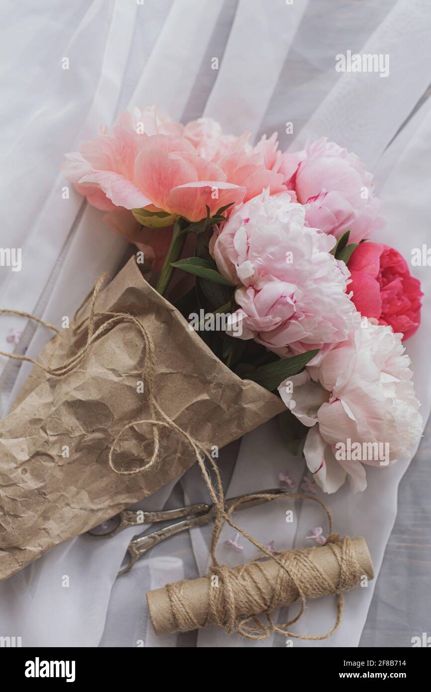 Beautiful stylish peony bouquet in paper, twine, scissors on soft fabric on dark wooden background, top view. Happy Mothers day. Pink and white peonie Stock Photo