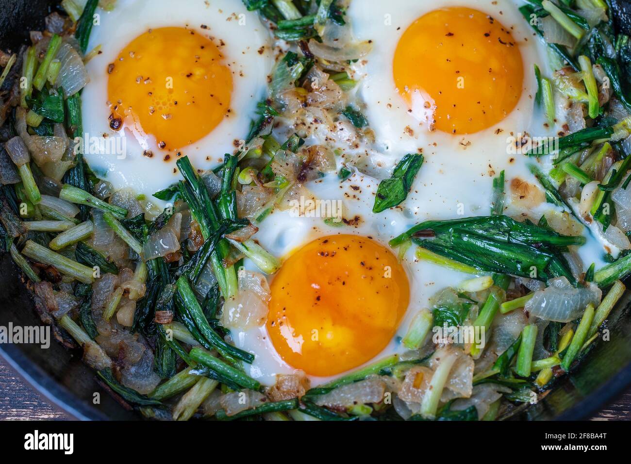 Shakshuka, fried eggs with green wild garlic leaves, onion, pepper and spices in cast iron pan, close up. Fried eggs with wild garlic as a shakshuka Stock Photo