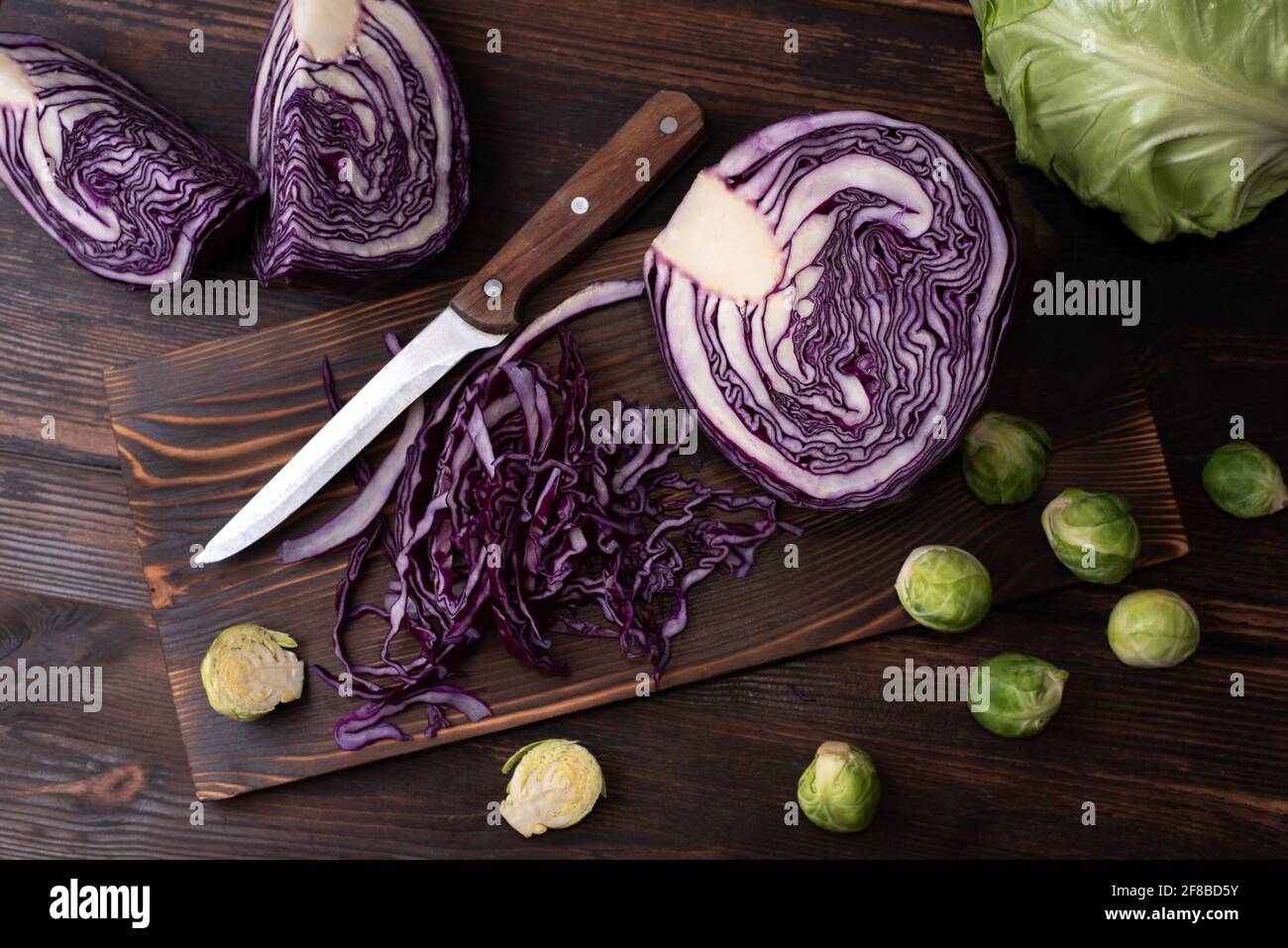 Chopped red cabbage on a cutting board on a wooden dark background with early cabbage head, brussels sprouts, rustic style, close up. Stock Photo