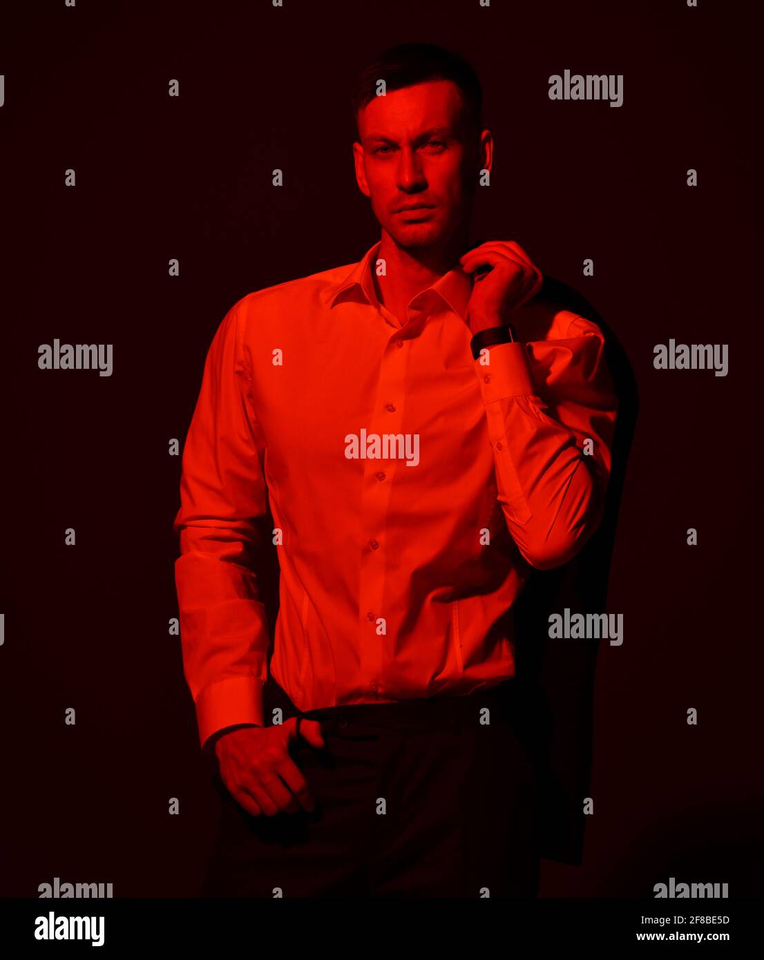 Young self-confident businessman yuppie in official suit stands holding jacket on his shoulder. Red light filter, gel Stock Photo