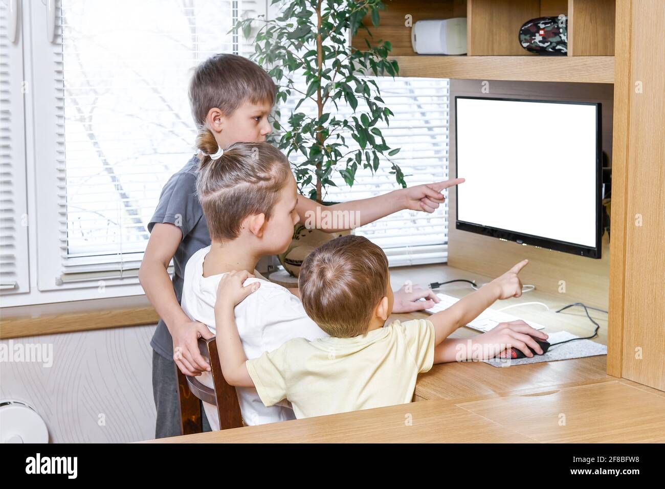 Schoolboys show empty computer white screen pointing with index fingers sitting and standing at wooden desk staying home at lockdown Stock Photo