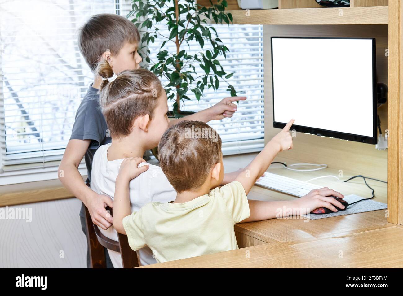 Schoolboys show empty computer white screen pointing with index fingers sitting and standing at wooden desk staying home at lockdown Stock Photo
