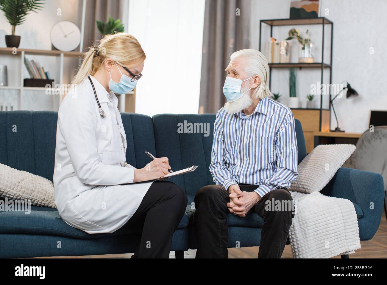 Mature woman doctor filling checkup form during home visit of retired bearded man patient, writing in documents, medical history or anamnesis, medical insurance contract. Stock Photo