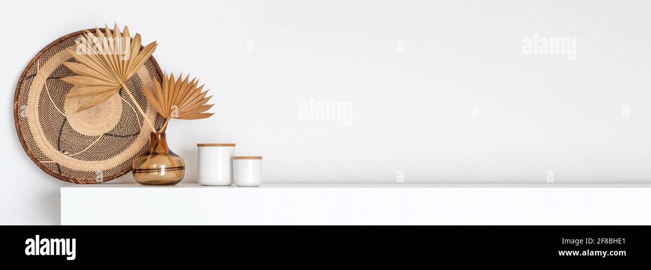 Header with coastal boho style interior decor, a brown glass vase with dried palm fans and a woven wall basket isolated on a white shelf Stock Photo