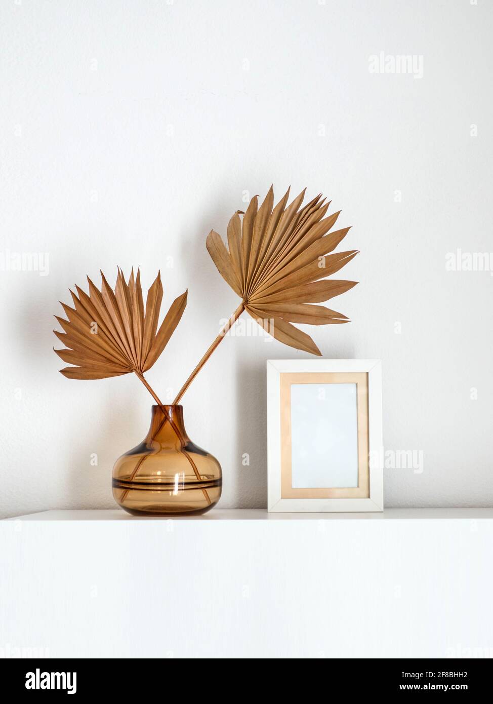 Empty white photo frame next to a glass vase with dried palm leaves. Stock Photo