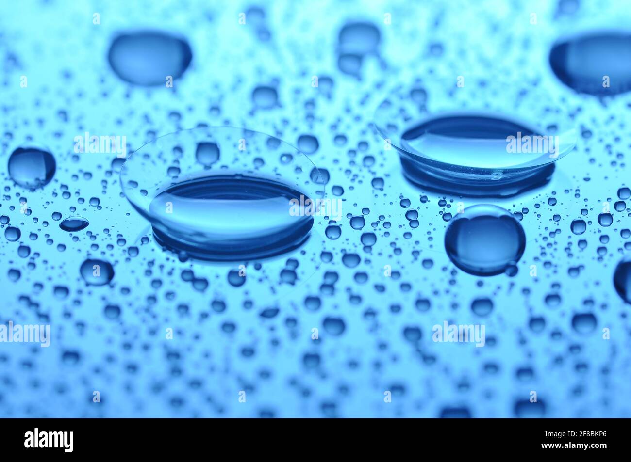 Two contact lenses with drops on light background. Blue toned image. Focus on long-distance lens. Concept sight. Stock Photo
