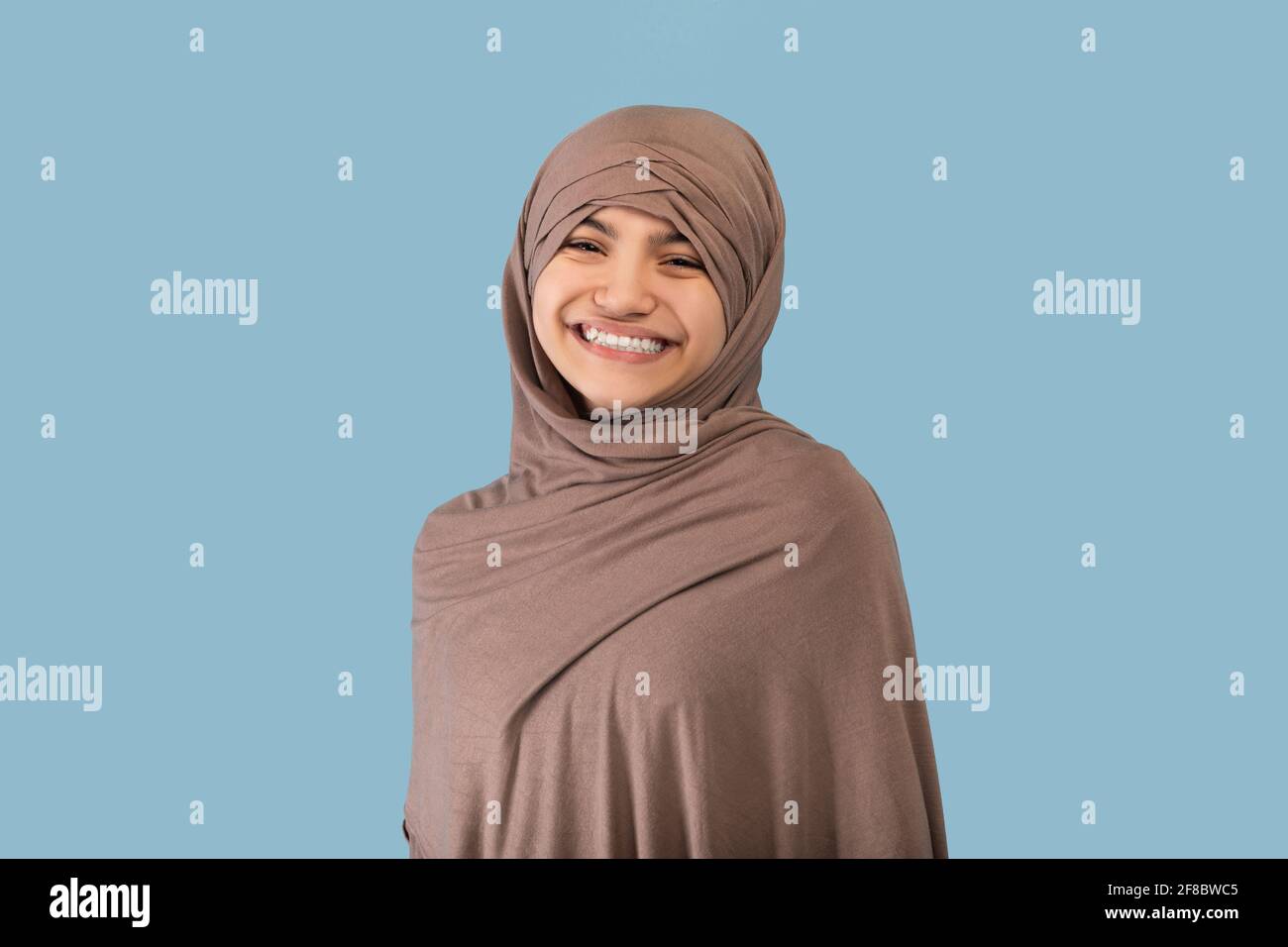 Portrait of positive Muslim teen girl in hijab smiling at camera over blue studio background Stock Photo