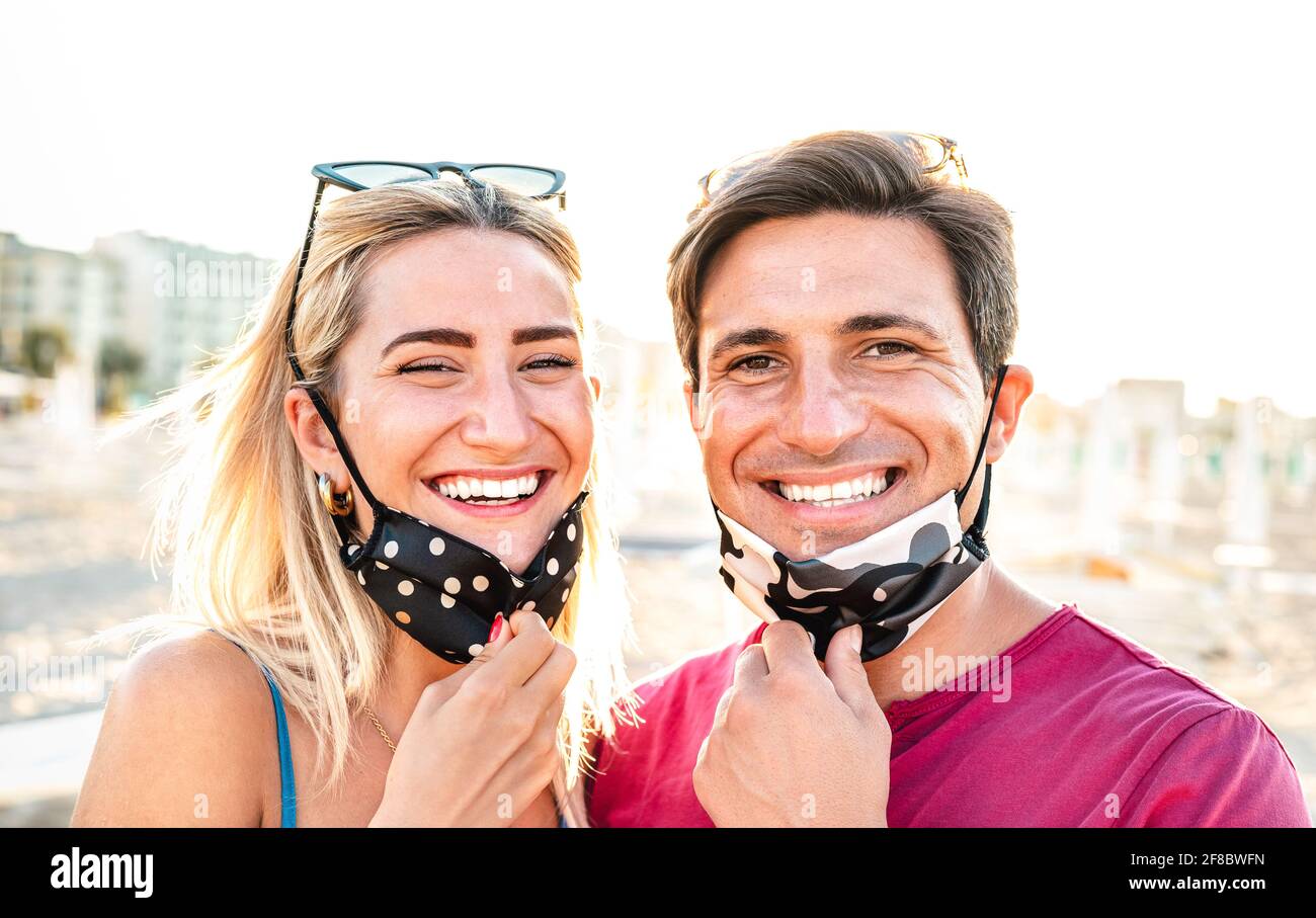 Young couple in love smiling with open face mask - New normal life style and relationship concept with happy lovers on positive mood at beach Stock Photo