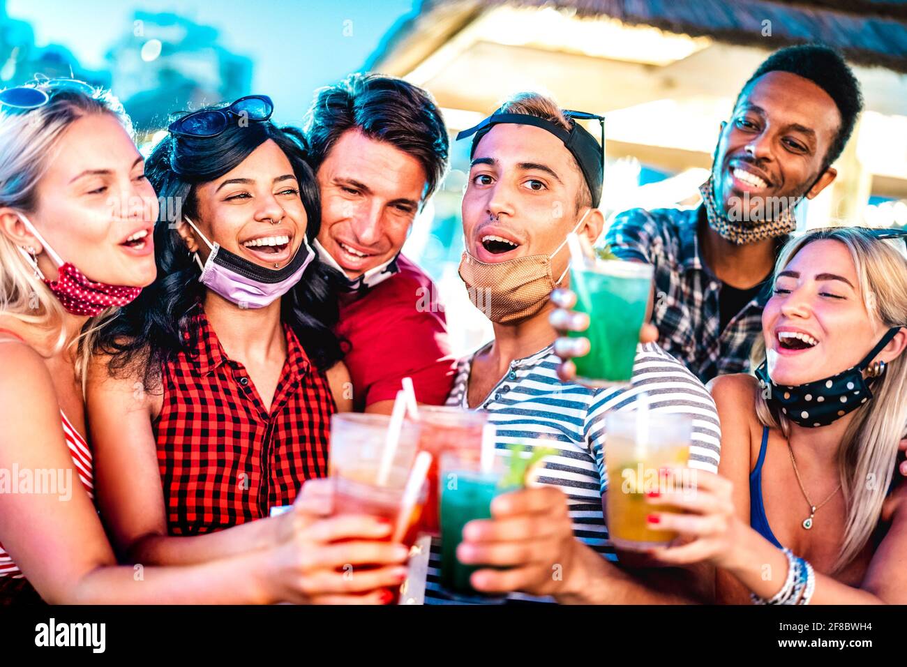Happy multicultural people toasting at night bar with open face masks - New normal life style concept with milenial friends having fun together Stock Photo
