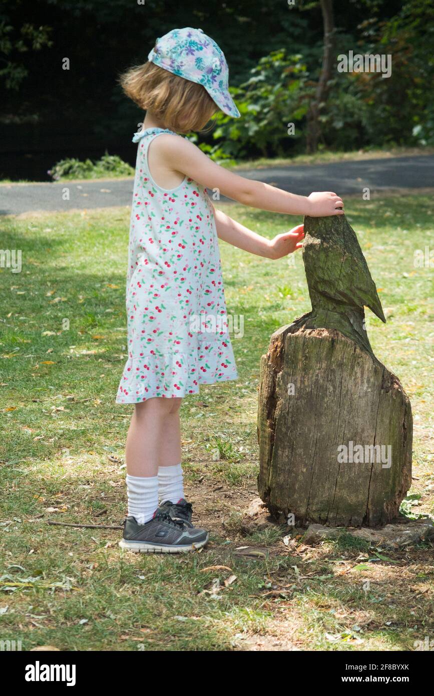 Young 5 year old girl aged five years with an owl ( or similar bird )  sculpture made from a sculptured tree stump in a public park. UK. (120) Stock Photo