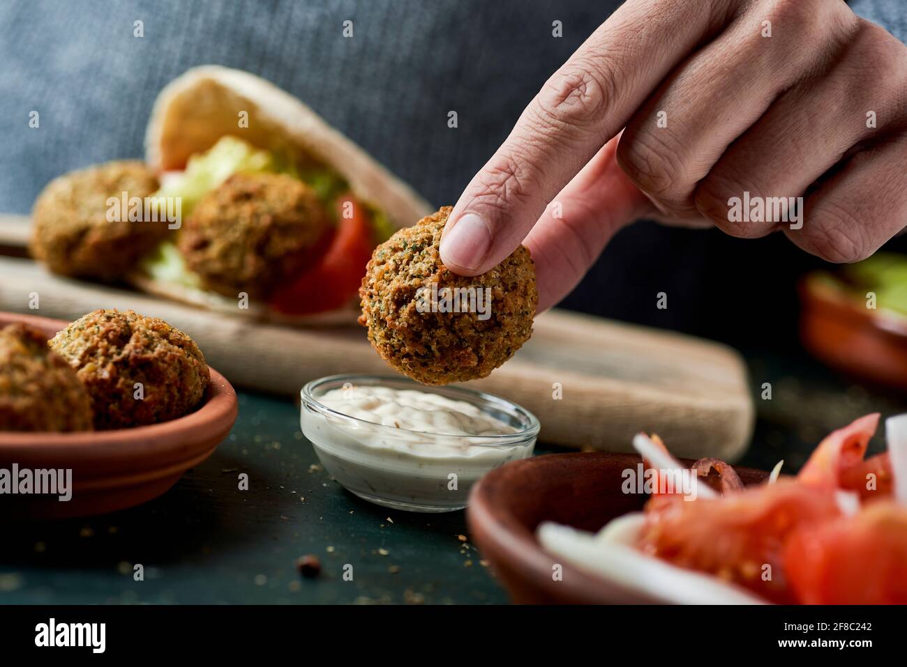 closeup of a young man dipping a falafel in a bowl of yogurt sauce sitting at a rustic green wooden table Stock Photo