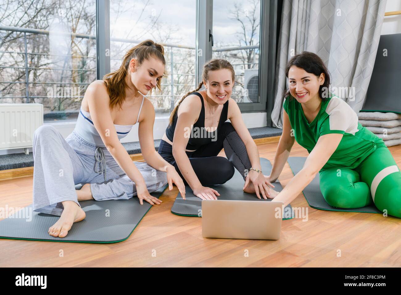Three sportive girls sit on mats in fitness studio during online yoga