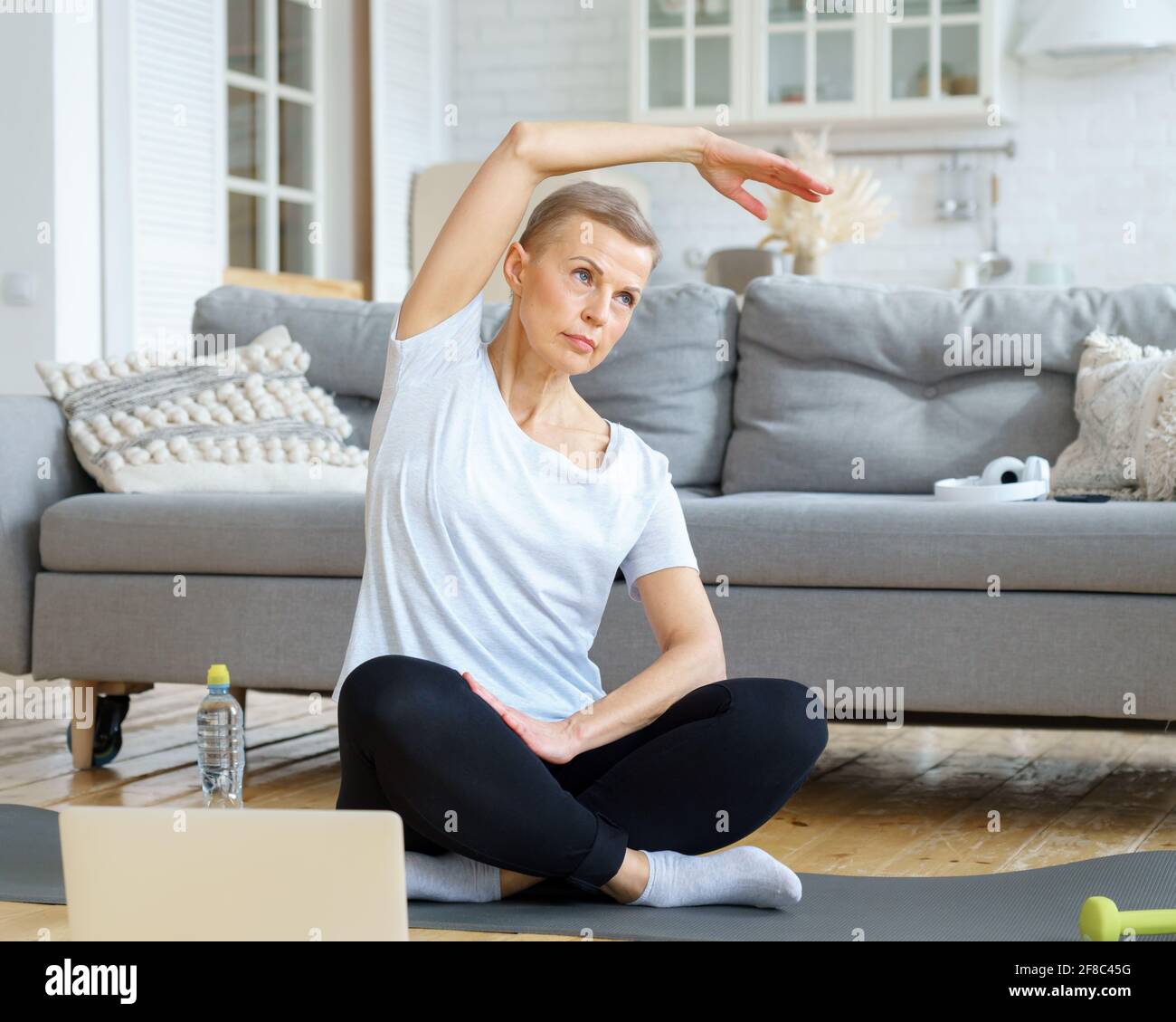 Senior woman stretching hand, online training with laptop in living room. Exercise for well-being, wellness for retired female. Domestic yoga practice. Flexible mature woman exercising on mat Stock Photo