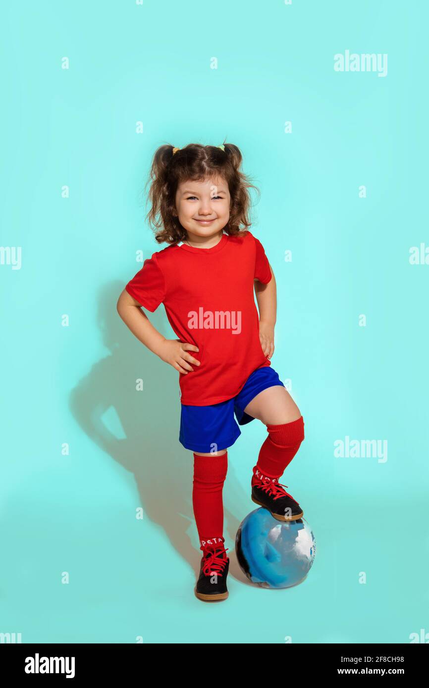 standing young soccer player girl 5-6 years holding football with foot on blue background Stock Photo