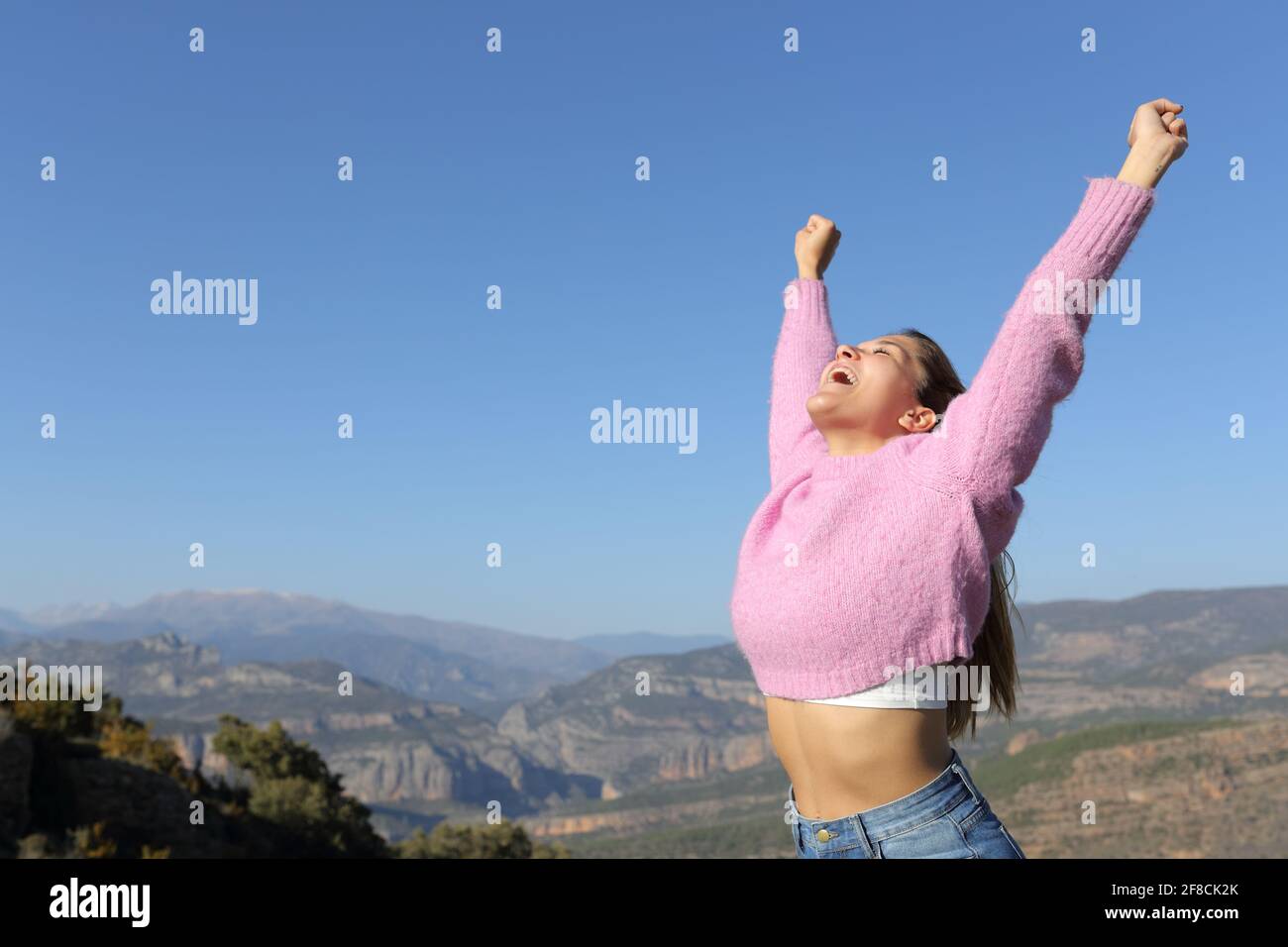 Excited woman in pink raising arms celebrating vacation in the mountain a sunny day Stock Photo