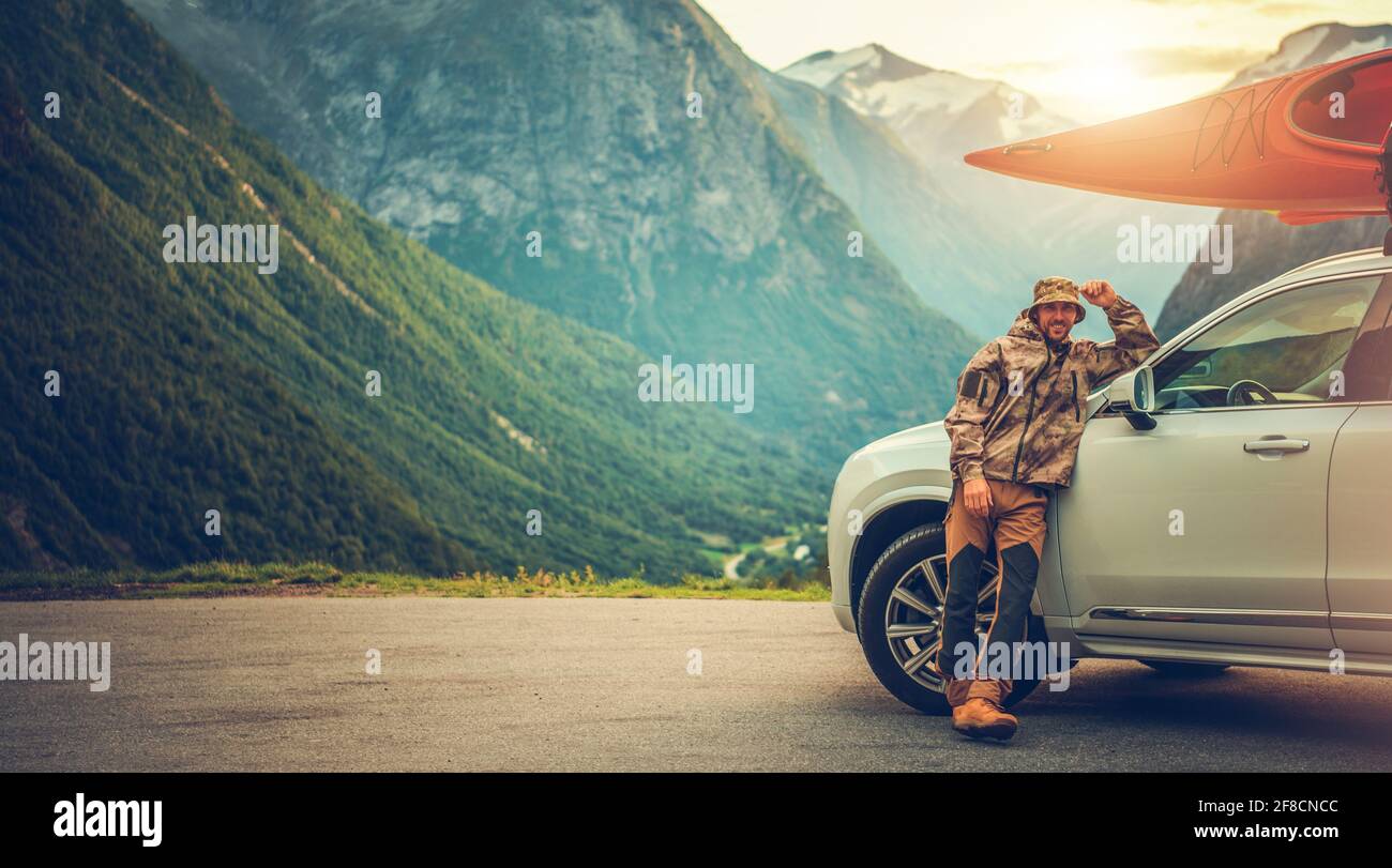 Caucasian Smiling Happy Men in His 40s on the Vacation Road Trip with Kayak on Roof of His Sport Utility Vehicle. Scenic Norwegian Landscape. Stock Photo