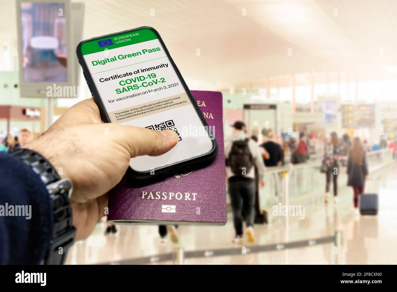 Male hand holding a phone with the European Union vaccination certificate on the screen and a traditional passport inside an airport. Immunity from Co Stock Photo