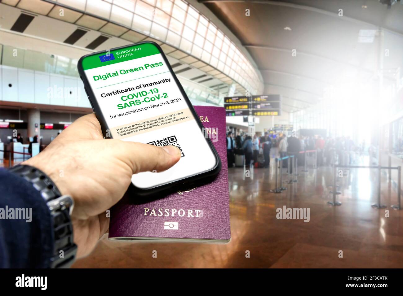 Male hand holding a phone with the European Union vaccination certificate on the screen and a traditional passport inside an airport. Immunity from Co Stock Photo